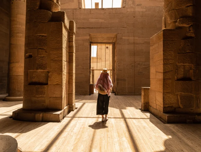 Person walking through a sunlit ancient Egyptian temple, surrounded by tall stone columns and intricate carvings.