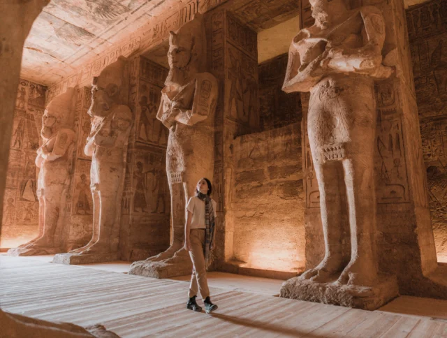 A person stands inside an ancient stone temple with towering pharaoh statues and intricate wall carvings.