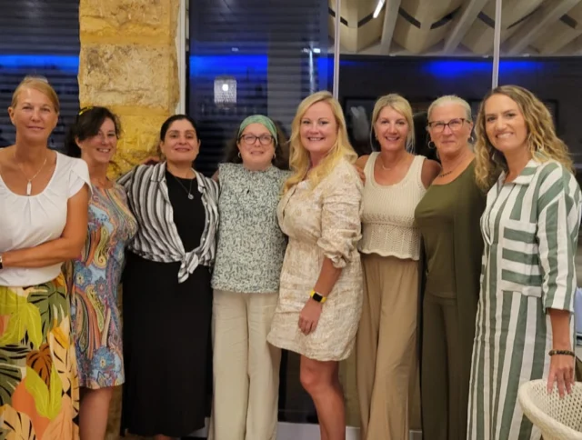 A group of nine women standing together indoors, smiling. They're dressed in a variety of casual and semi-formal outfits.