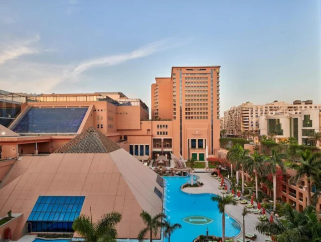 A hotel complex with pyramid-like structures, a central pool, and surrounding palm trees, set against a cityscape.