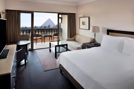 Hotel room with a large bed, a sofa, and a TV. The room features a balcony with chairs and a table, offering a view of a pyramid in the distance through glass doors.