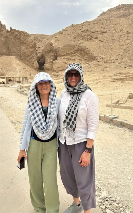 Two people in light clothing and headscarves stand on a pathway in a rocky, desert landscape.