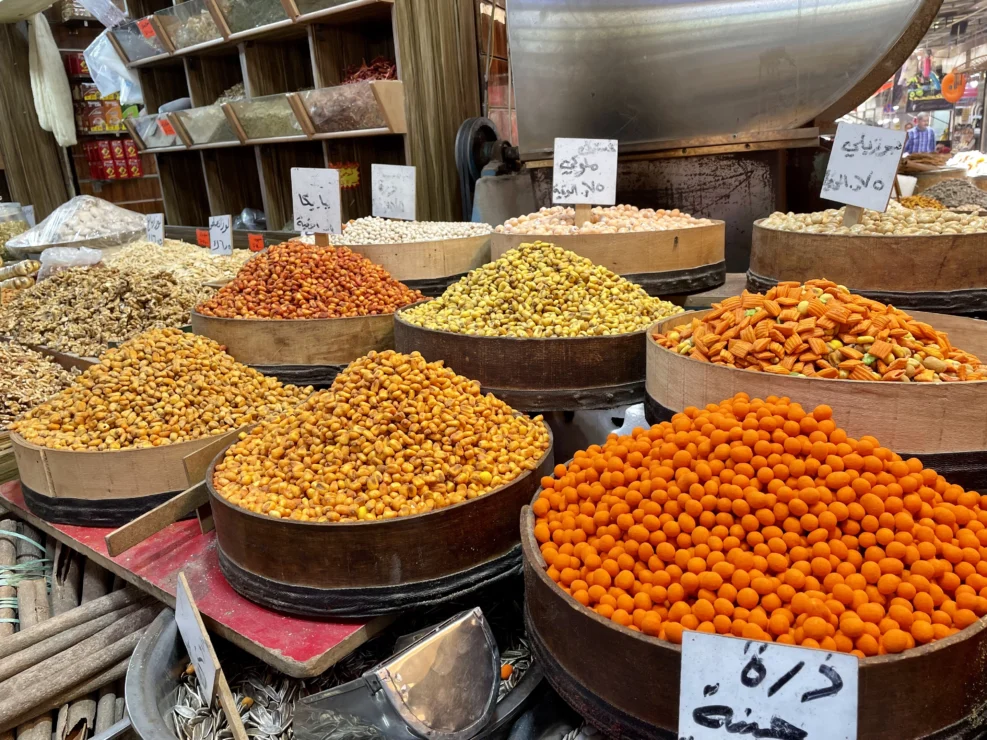 Various colorful spices and dried foods in round baskets at a market stall, each with handwritten labels.