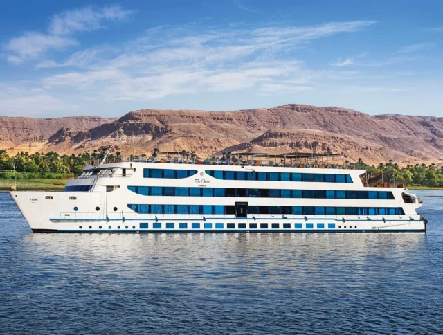 A large white and blue cruise ship sails on a river with arid, rocky hills and clear skies in the background.