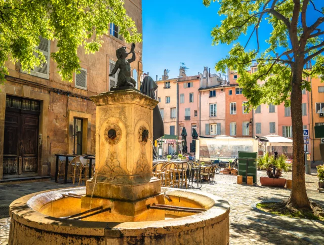 A sunlit plaza features a stone fountain with a statue, surrounded by trees, tables, and colorful buildings under a clear blue sky.