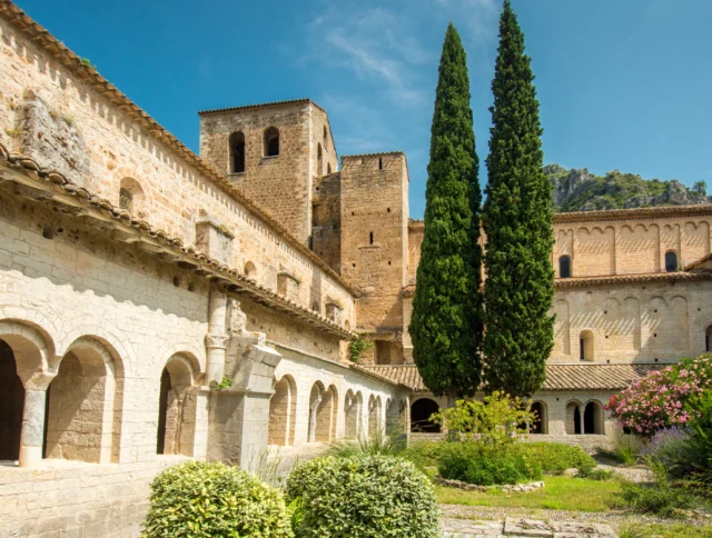 Historic stone monastery with arched walkways, two tall cypress trees, and a garden under a clear blue sky.