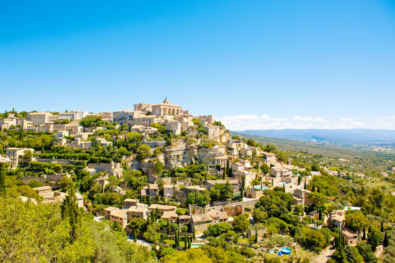 A hilltop village with stone buildings and lush greenery under a clear blue sky.