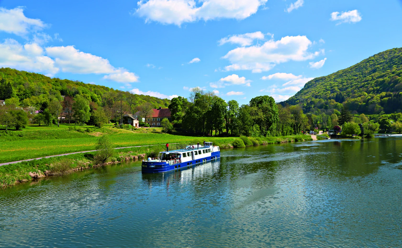 A blue and white boat sails along a calm river, surrounded by green hills and trees under a partly cloudy sky.