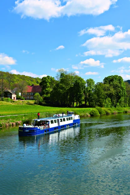 A blue and white boat sails along a calm river, surrounded by green hills and trees under a partly cloudy sky.