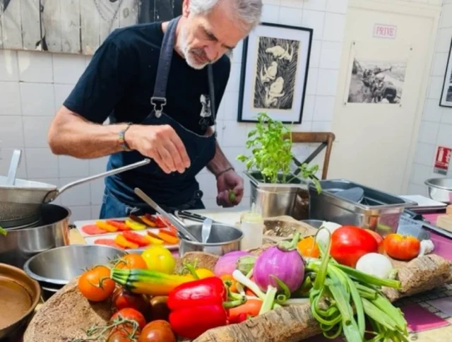 A chef prepares food in a kitchen with a wooden tray of colorful vegetables in the foreground.