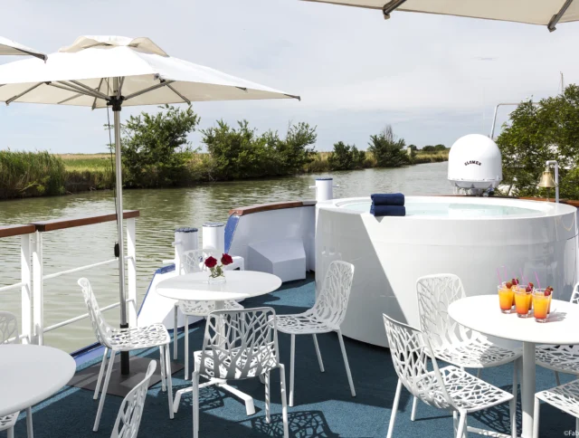 A boat deck features a round hot tub, white chairs and tables, and a blue floor. Umbrellas provide shade near a river with green vegetation.