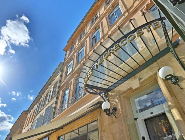 Street view of a stone building with a sign reading "Le Negrecoste" and a glass awning. The sky is clear with a few clouds and the sun shining brightly.