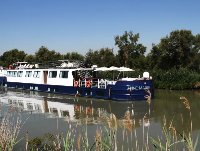 A blue and white barge cruises on a calm river, surrounded by greenery and under a clear blue sky.