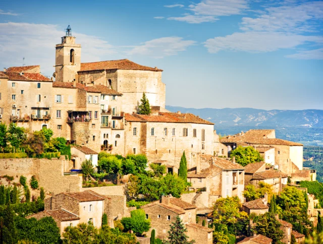 A scenic view of a hilltop village with rustic stone buildings and a church, surrounded by trees, overlooking a vast landscape under a partly cloudy sky.