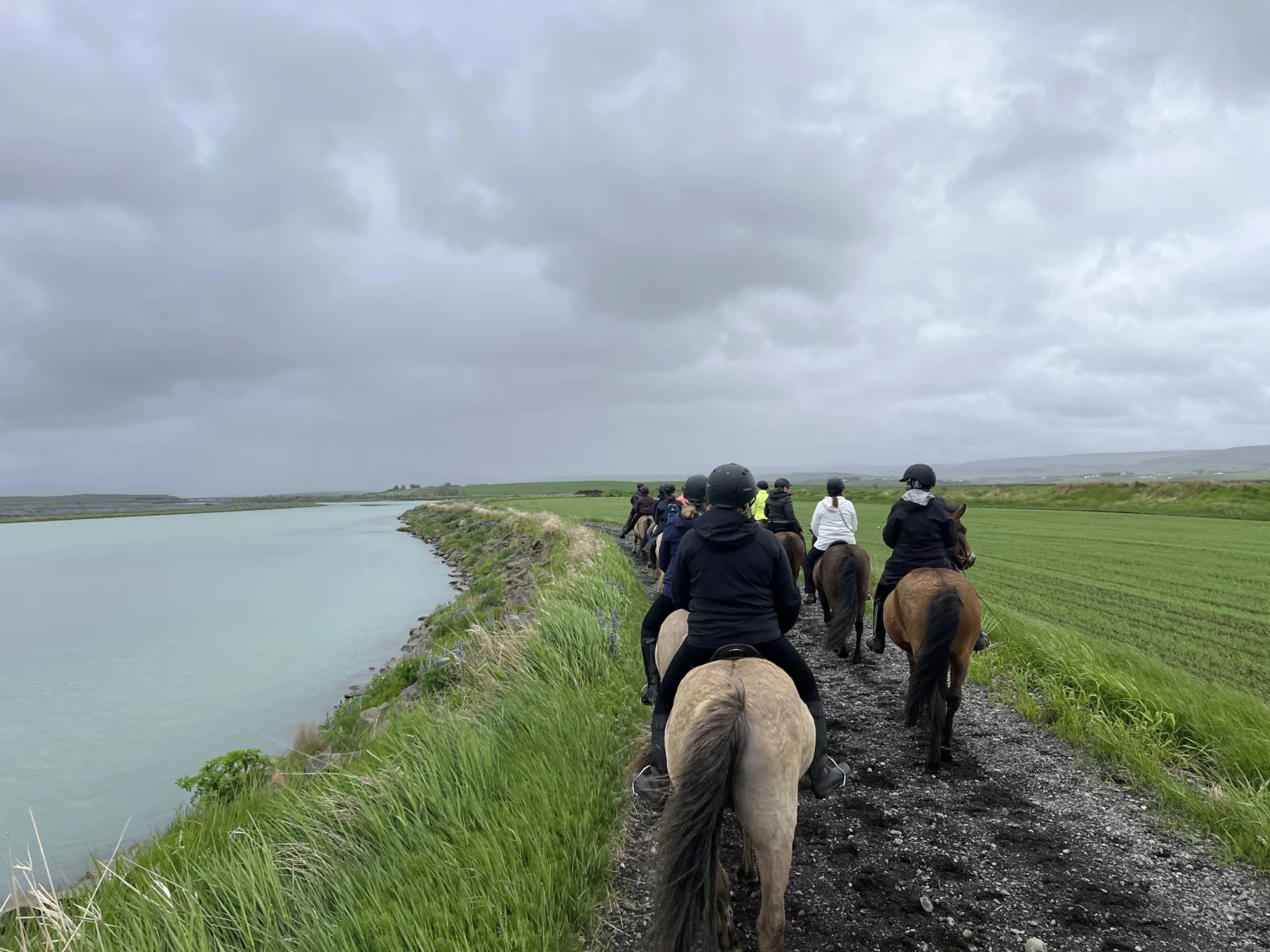 A group of people on horseback rides along a dirt path beside a body of water and a grassy field under a cloudy sky.