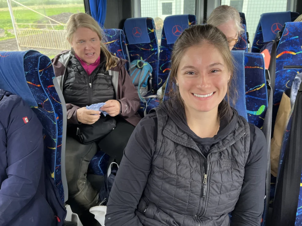 Two women seated on a bus. One is smiling at the camera, while the other is knitting. The bus seats are blue with a colorful pattern.