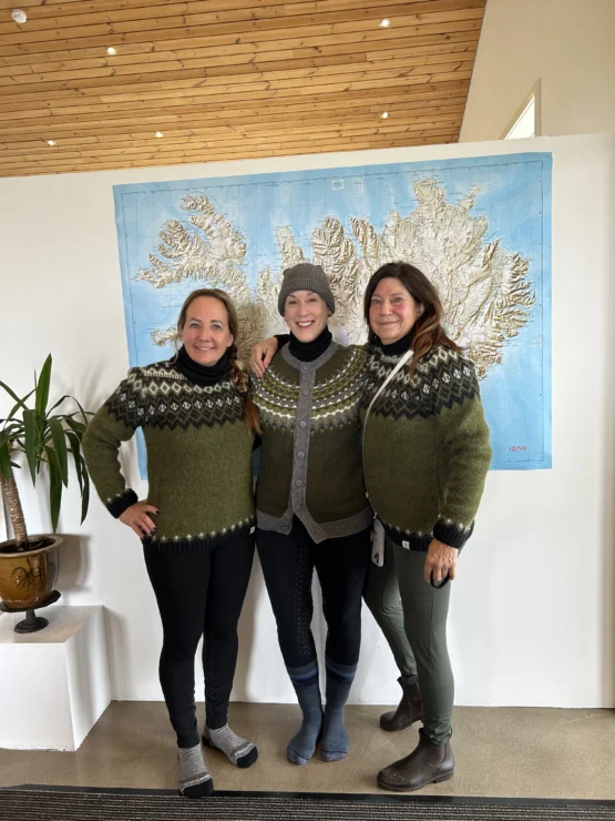 Three people in knitted sweaters stand together smiling in front of a large wall map.