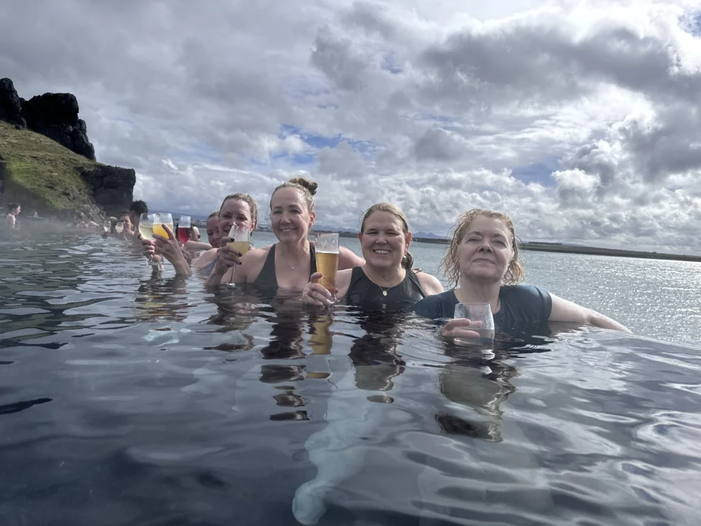 Four people in a hot spring, each holding a beverage, with a cloudy sky and rocky landscape in the background.