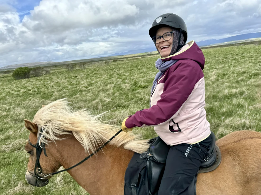 A person wearing a helmet smiles while riding a brown horse across an open grassy field under a cloudy sky.