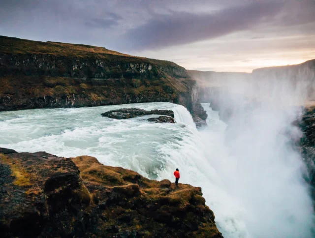 A person in a red jacket stands on a rocky cliff overlooking a large, misty waterfall under a cloudy sky.