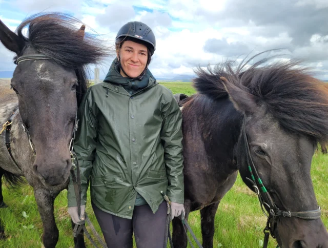 A person in a green jacket and helmet stands between two dark-haired horses in a grassy field.