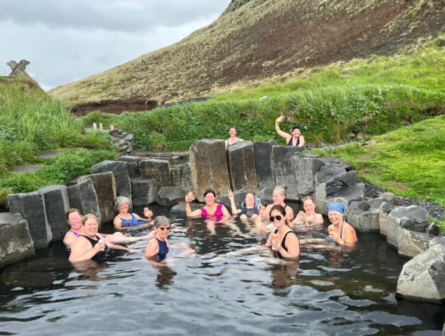 A group of people relaxing in a natural hot spring surrounded by greenery and rocky hills.