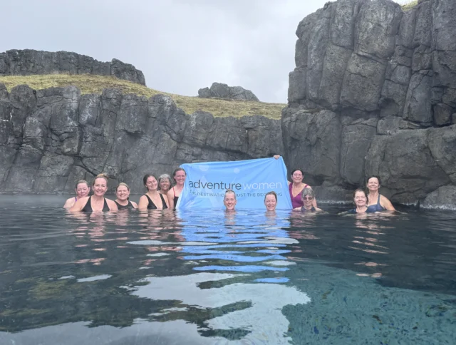 A group of women in swimsuits stand in a hot spring, holding a blue "adventure women" banner, surrounded by rocky cliffs.