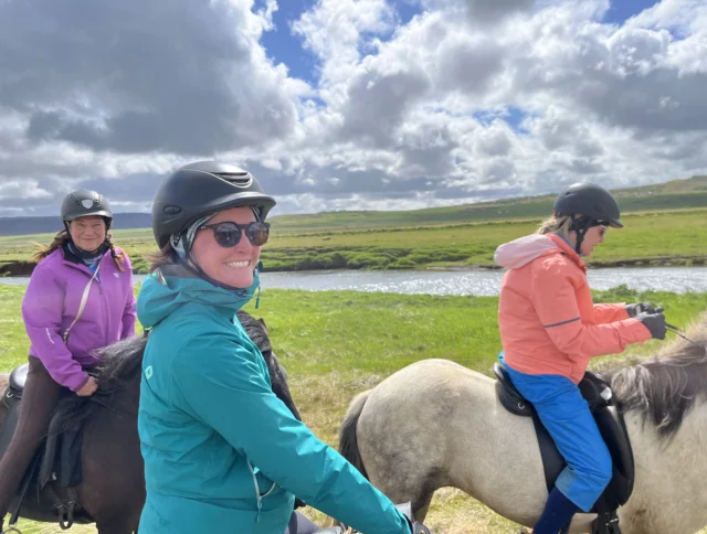 Three people in helmets ride horses alongside a river on a sunny day with clouds in the sky.