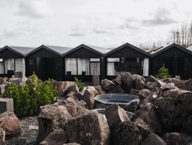 Row of black cabins with large windows, set against a cloudy sky. Foreground features scattered rocks and sparse greenery.