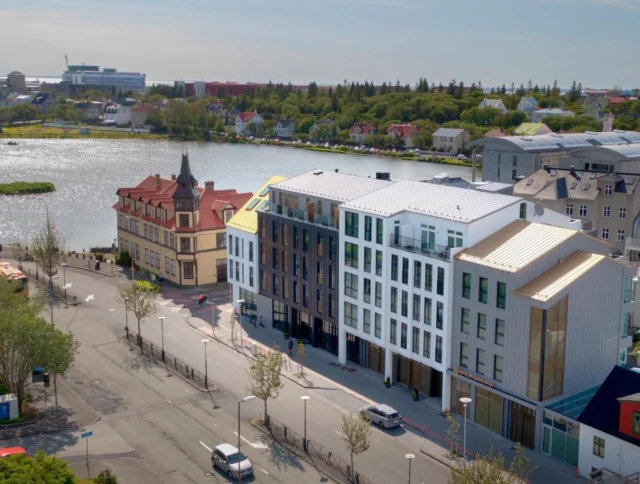 Aerial view of a street with modern and historic buildings near a lake, surrounded by greenery and a cityscape in the background.