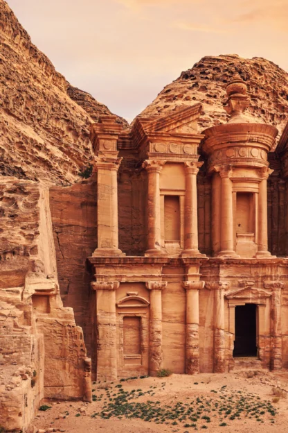 The image shows the ancient rock-cut architecture of Al Deir, the Monastery, in Petra, Jordan, with a mountainous backdrop and an orange sky.