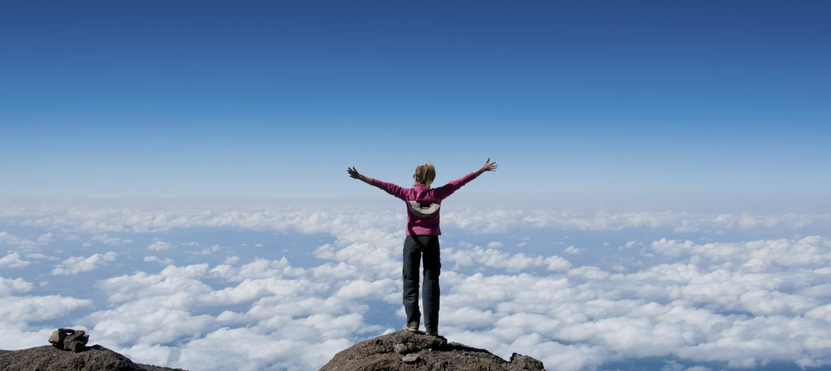 Person standing on a rocky peak with arms outstretched, overlooking a sea of clouds under a clear blue sky.