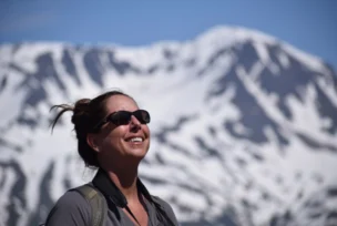 Woman smiling and wearing sunglasses, standing outdoors with a snowy mountain in the background.