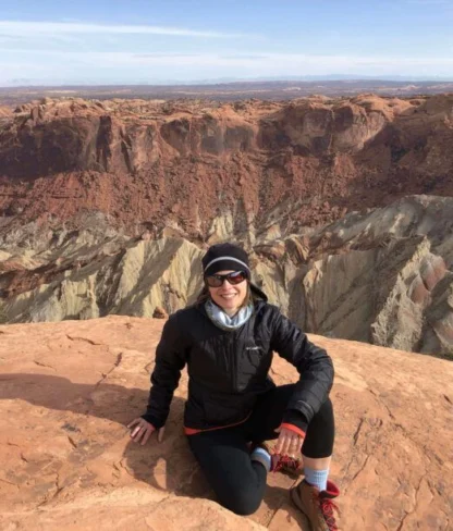 Person sitting on a rocky cliff with a layered canyon landscape in the background, wearing a jacket and hat.