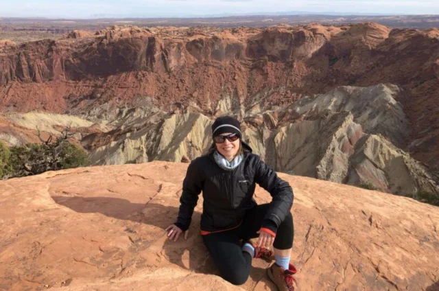 Person sitting on a rocky cliff with a layered canyon landscape in the background, wearing a jacket and hat.