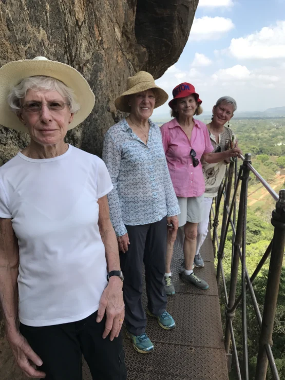 Four women wearing hats stand on a metal walkway by a rock formation with a scenic view in the background.