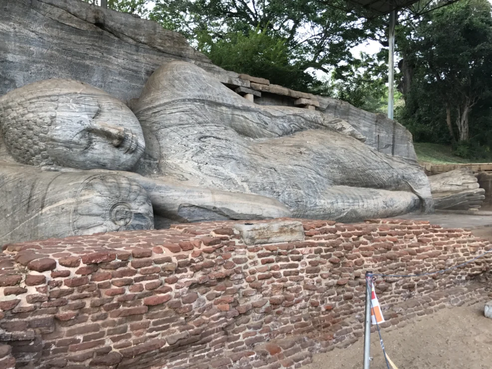 A large stone statue of a reclining Buddha is carved into a rock face, surrounded by trees and brickwork in a serene outdoor setting.