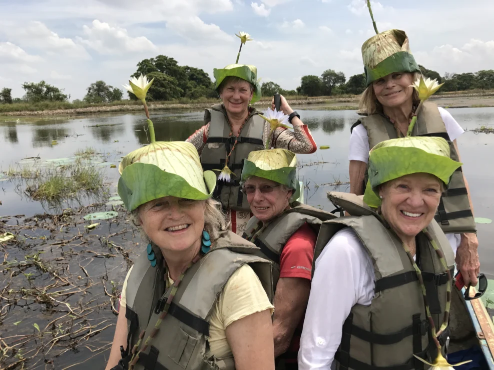 Five women wearing life vests and leaf hats with flowers smile while sitting in a boat on a calm waterway.