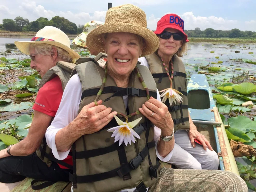 Three women in life vests and sun hats are seated in a boat on a lily-covered lake, smiling at the camera.