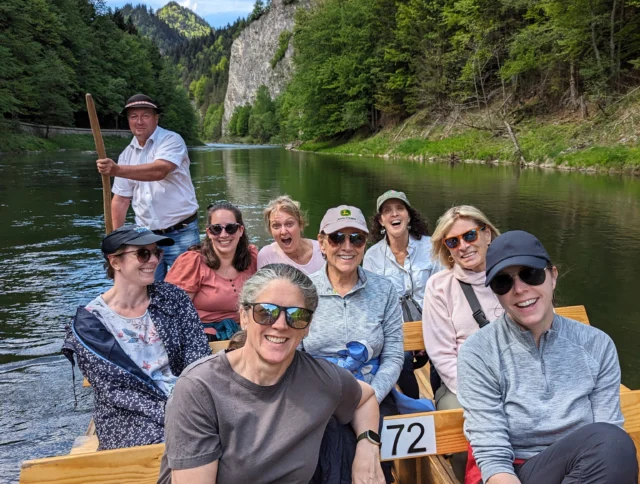 A group of people sitting in a wooden boat on a river surrounded by lush, forested cliffs. One person is standing at the back steering with a pole. It's a sunny day.