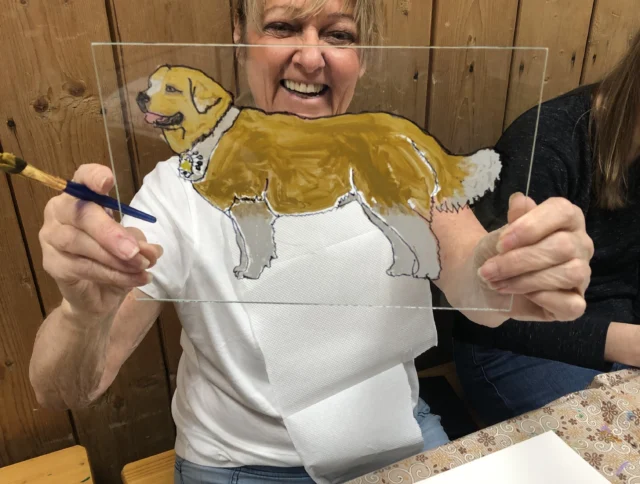 A woman holds up a painted glass pane depicting a golden dog. She is seated at a table with art supplies.