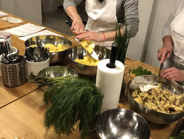 A person in a kitchen chops potatoes next to bowls of herbs and greens on a wooden counter.