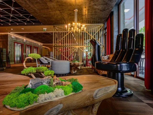 Modern interior with a large black hand-shaped chair, wooden table with greenery, and artistic lighting fixtures in a spacious room.