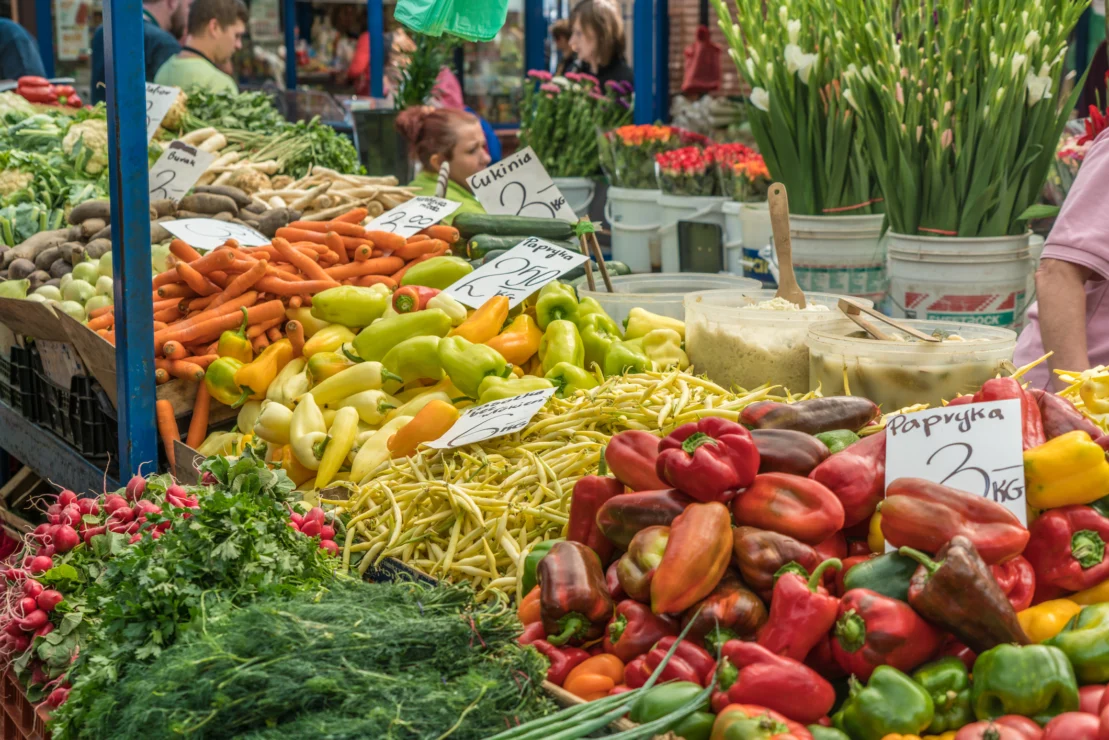 A vibrant market stall displays various fresh vegetables, including peppers, carrots, and herbs, with price signs in a lively outdoor setting.