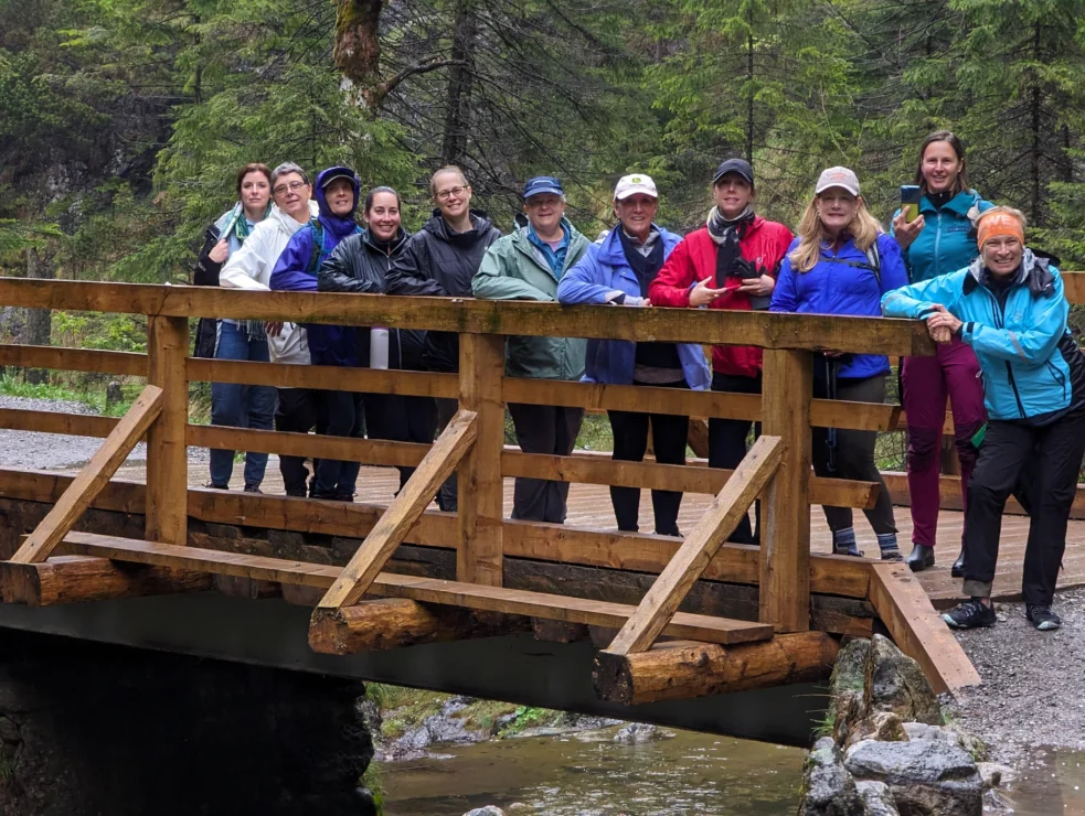 A group of twelve people in outdoor clothing stands on a wooden bridge over a stream in a forested area.