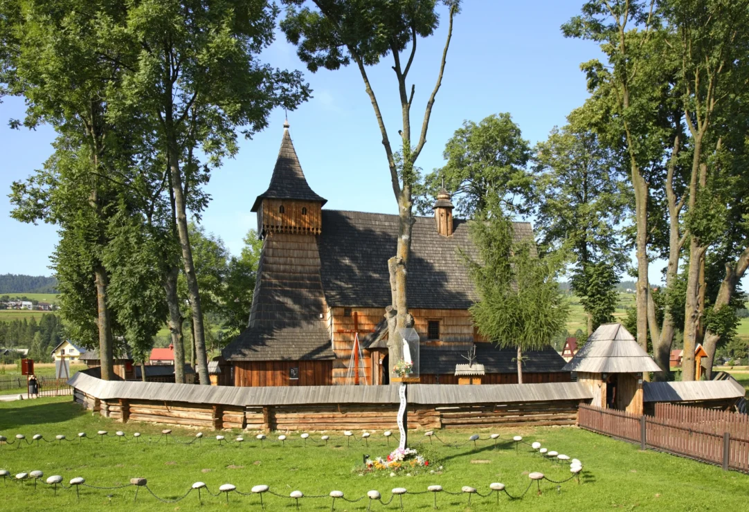 Wooden church with a sloped roof surrounded by a wooden fence and trees, set in a grassy area with a row of stones arranged in a circle.