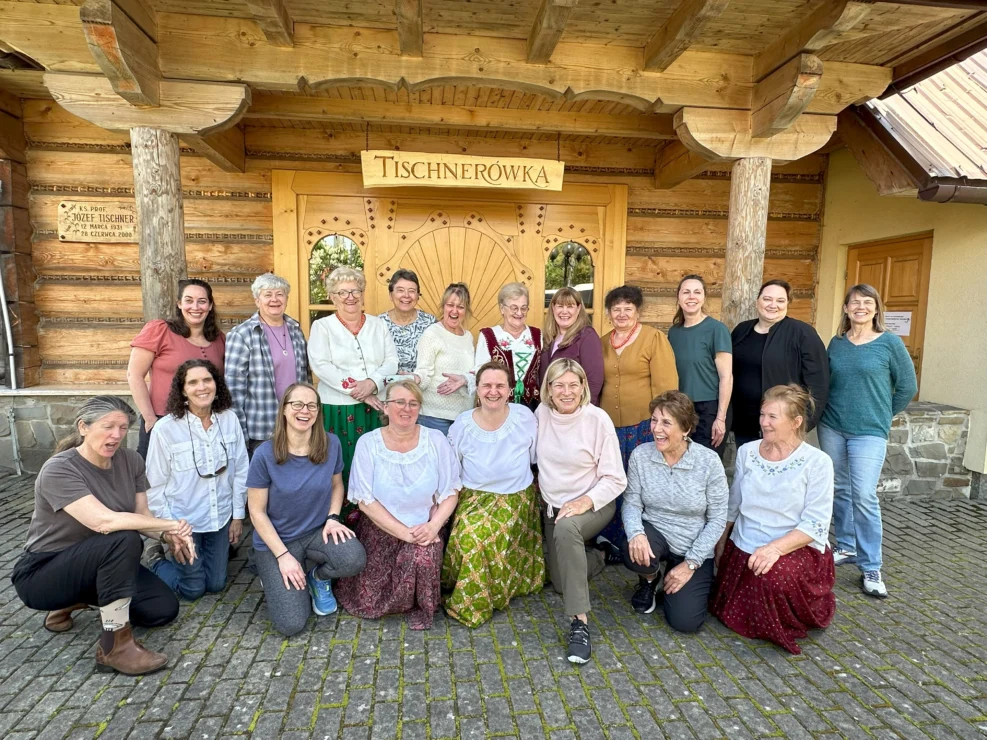 A group of people smiling and posing in front of a wooden building with a sign that reads "Tischnerówka.