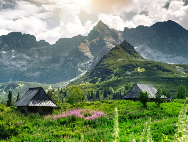 Two wooden huts sit in a lush green meadow with pink flowers, framed by tall, rugged mountains and a partly cloudy sky.