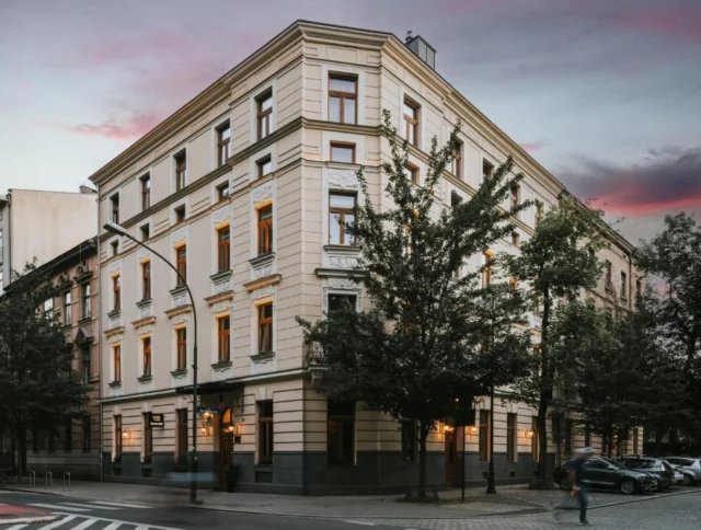 A four-story corner building with illuminated windows, set against a sunset sky. Trees partially obscure the facade.