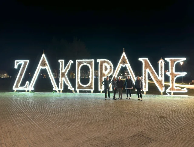 People standing in front of an illuminated sign spelling "ZAKOPANE" at night.